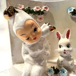 May include: A white ceramic figurine featuring a winking cherub with gold accents, a white bunny, and floral details. The cherub is wearing a white outfit with a gold-colored headpiece. The bunny has pink-lined ears and a red mouth. Pink and blue flowers are also present.