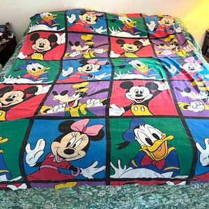 May include: A bedspread featuring a grid pattern of colorful squares, each displaying a classic cartoon character including Mickey Mouse, Minnie Mouse, Donald Duck, and Goofy. The design uses vibrant red, blue, green, and purple background colors.