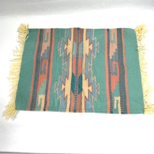 May include: A rectangular woven rug featuring a geometric pattern in shades of teal, terracotta, tan, and dark gray. The rug is finished with cream-colored fringe on the left and right edges, creating a textured border along the sides of the piece.