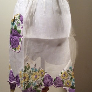 May include: White apron with a purple and yellow floral print. The apron has a ruffled waistband and a large pocket on the front.