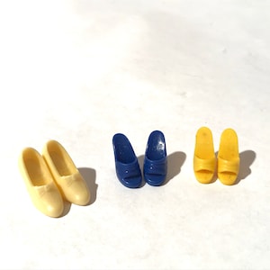 May include: Three pairs of doll shoes. One pair is yellow, one pair is blue, and one pair is cream.