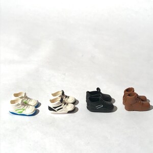 Op de afbeelding: Vier paar miniatuurschoenen, waaronder witte sneakers met groene accenten, zwarte sneakers, witte sneakers met zwarte accenten en bruine laarzen.