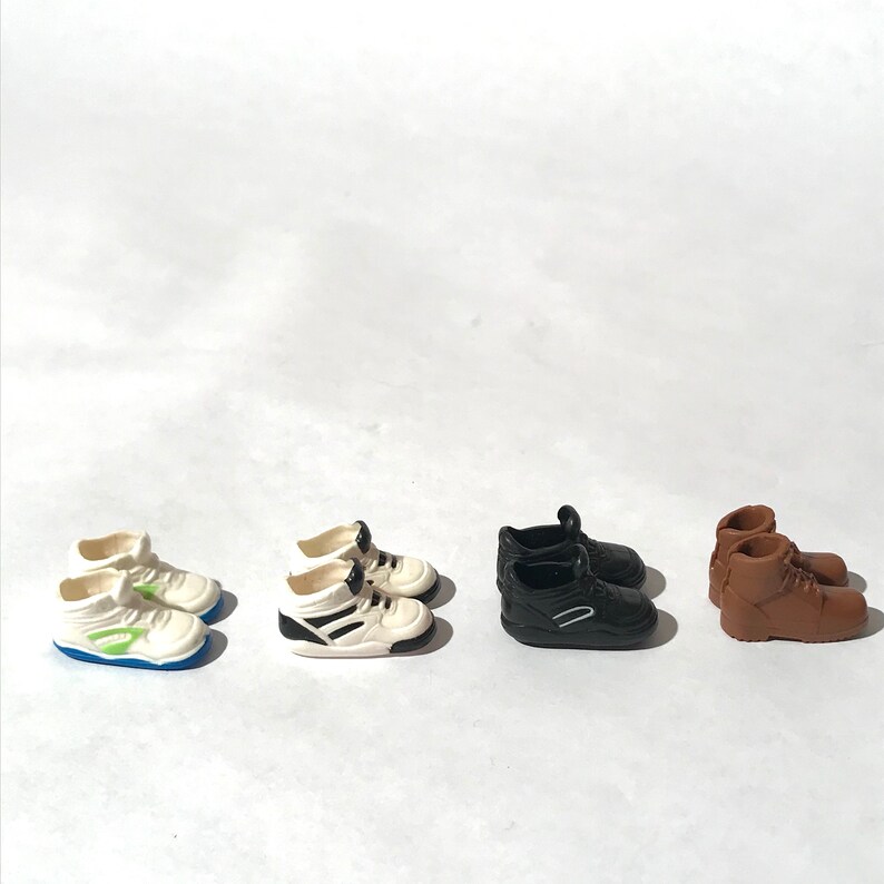 Op de afbeelding: Vier paar miniatuurschoenen, waaronder witte sneakers met groene accenten, zwarte sneakers, witte sneakers met zwarte accenten en bruine laarzen.