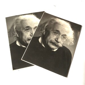 May include: Two black and white photographs of Albert Einstein. The photographs show Einstein with his signature white hair, mustache, and dark sweater. The images are slightly overlapping, with the edges of the photos visible.