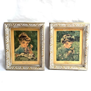 May include: Two framed vintage prints. One print features a girl with a cat and the text "Little Boynoles". The other print shows a girl with a butterfly and the text "Summer's Victor". Both prints are in ornate white and gold frames.