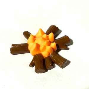 May include: A plastic toy campfire with brown logs and an orange flame.