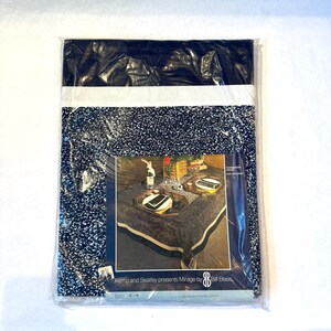May include: A black and white tablecloth with a white border. The tablecloth is packaged in plastic and features a label that reads "Kemp and Beatley presents Mirage by Bill Blass". The label also includes the size of the tablecloth, 83 x 96 inches.