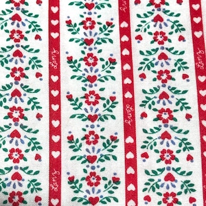 May include: A white fabric featuring vertical red stripes alternating with floral patterns. The red stripes contain a repeating white heart motif and the text Lanz. The floral columns consist of red flowers, green leaves, and small blue dots.