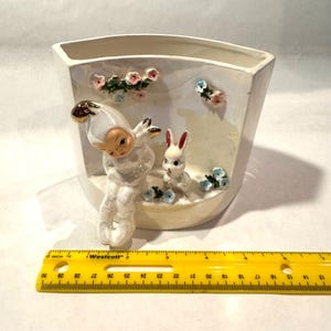 May include: A vintage ceramic planter with a whimsical design. It features a seated figure with a gold-tipped hat and a white rabbit, surrounded by floral accents. The planter is approximately 6 inches wide.