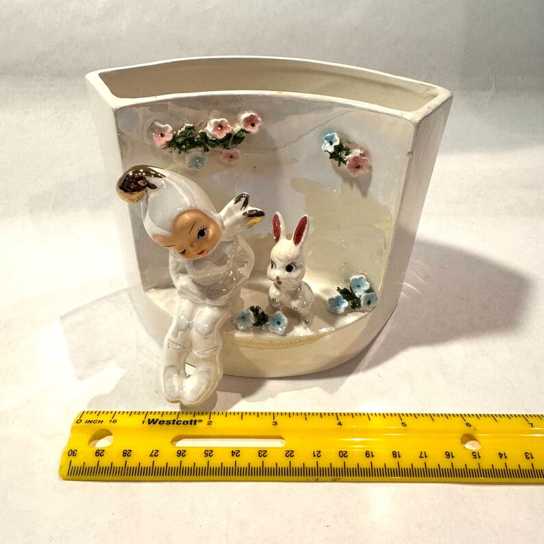 May include: A vintage ceramic planter with a whimsical design. It features a seated figure with a gold-tipped hat and a white rabbit, surrounded by floral accents. The planter is approximately 6 inches wide.