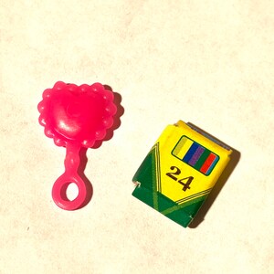May include: A pink plastic heart-shaped rattle with a handle and a box of 24 crayons.