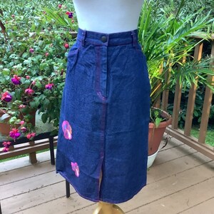 May include: A dark blue denim skirt with a pink and purple floral design. The skirt has a front slit and a button closure.