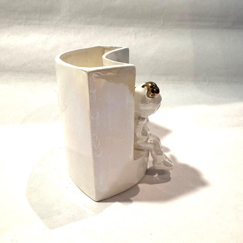 May include: A white ceramic vase with a unique design, featuring a sitting frog figurine with gold-colored eyes. The vase has a curved opening and a rectangular base, creating a modern and whimsical aesthetic.