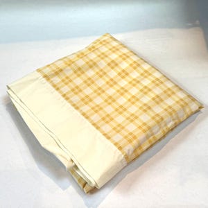 May include: Folded yellow and white plaid pillowcase with a cream-colored border. The pillowcase features a classic plaid pattern in shades of yellow and white, with a smooth, cream-colored border.