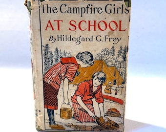 Op de afbeelding: De omslag van het boek The Campfire Girls At School van Hildegarde G. Frey toont een illustratie van twee meisjes in rood-wit gestreepte shirts en rode hoeden, die kamperen in een bosrijke omgeving. De meisjes wassen afwas in de buurt van een tent en een kano.