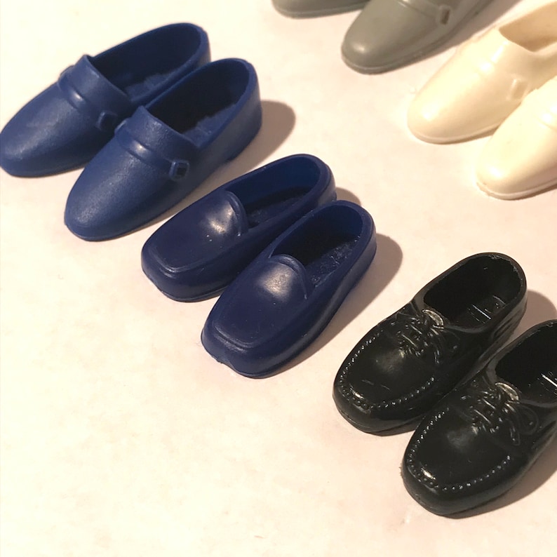 May include: Six pairs of plastic doll shoes in various colours and styles. The shoes include black loafers, black lace-up shoes, blue loafers, white loafers, and grey loafers.