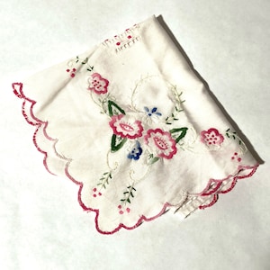 May include: White embroidered fabric doily with pink and blue flowers and green leaves. The doily has a scalloped edge.