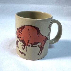 May include: A beige ceramic mug featuring a dark reddish-brown graphic illustration of a large bison, accompanied by smaller animal figures. The mug has a cylindrical body and a curved handle on the right side, with a smooth, glossy finish throughout.