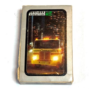 May include: A vintage video game cartridge featuring a semi-truck with bright headlights and the text "ROAD XPEDITOR 2". The game cartridge has a silver and black border.