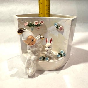 May include: A white ceramic planter with a whimsical design. It features a seated figure with a gold-accented hat and a white rabbit. Pink and blue floral accents decorate the planter. The planter measures approximately 7 inches tall.