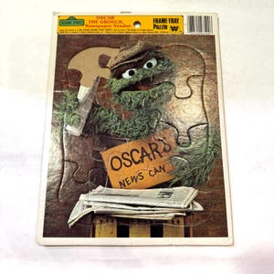 May include: A vintage Sesame Street frame-tray puzzle featuring Oscar the Grouch as a newspaper vendor. The puzzle shows Oscar holding a bottle and a sign that reads "OSCAR'S NEWS CAN". The puzzle is made of cardboard and has a white border.