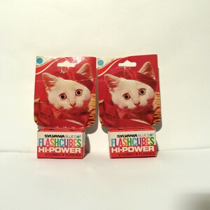 May include: Two boxes of Sylvania Blue Dot Flashcubes Hi-Power flashbulbs. Each box contains 2 cubes and 8 flashes. The boxes are red and white with a picture of a white cat wearing a red hat.