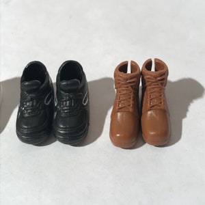 Op de afbeelding: Twee paar poppen schoenen. Een paar is zwarte sneakers met witte accenten. Het andere paar is bruine leren laarzen met veters.