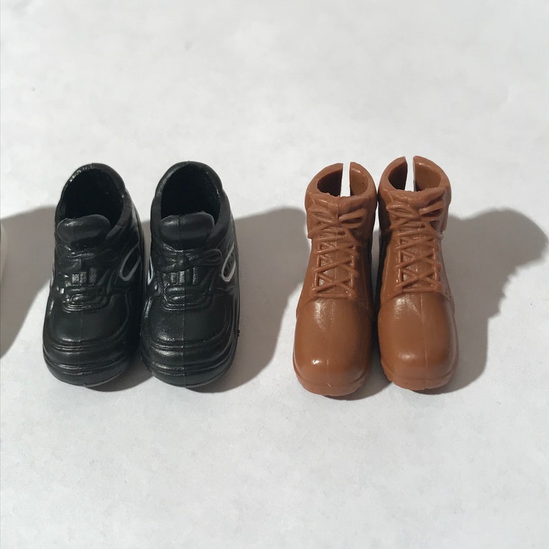 Op de afbeelding: Twee paar poppen schoenen. Een paar is zwarte sneakers met witte accenten. Het andere paar is bruine leren laarzen met veters.