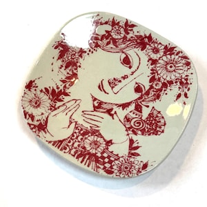 May include: A square ceramic plate with a light green background and a red illustration. The illustration depicts a woman's face and hands surrounded by floral patterns. The plate has a glossy finish.