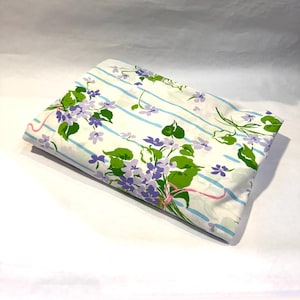 May include: Folded white sheet with a floral pattern. The sheet features light blue stripes, purple flowers, green leaves, and pink ribbon accents. The floral design is spread across the sheet.