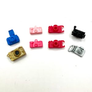 May include: Assortment of miniature plastic camera charms in various colors, including blue, pink, gold, black, and silver. The charms are designed to look like vintage cameras.