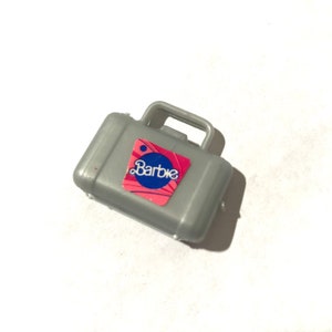 May include: A gray plastic suitcase with a handle and a pink and blue Barbie logo.