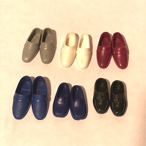 May include: Six pairs of doll shoes in various colours, including grey, white, burgundy, blue, and black. The shoes are all loafers with a simple design.
