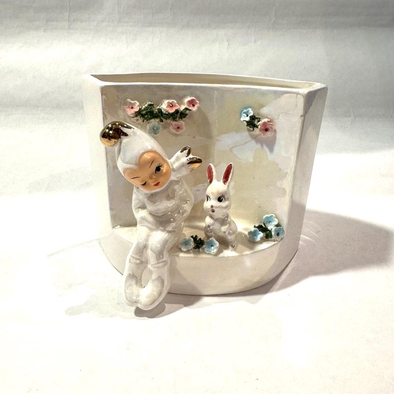 May include: A white ceramic planter with a whimsical design. It features a seated figure with a gold-tipped hat and a white rabbit. Pink and blue floral accents decorate the planter's interior.