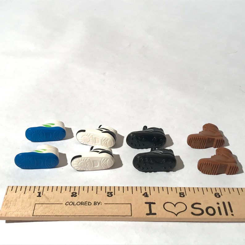 Op de afbeelding: Zes paar miniatuur plastic schoenen in verschillende kleuren, waaronder wit, zwart, blauw en bruin. De schoenen zijn gerangschikt in twee rijen van drie, met een liniaal op de voorgrond. De liniaal heeft de tekst "I Love Soil!" erop gedrukt.