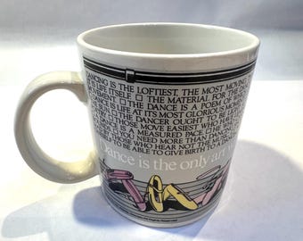 May include: White ceramic mug with a curved handle. The mug features text about dance and illustrations of ballet shoes in pink and yellow. The text includes the phrase "Dance is the only art".