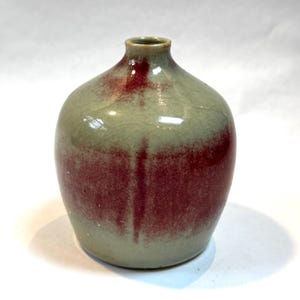 May include: A small, round ceramic vase with a narrow neck. The vase has a light green base with a reddish-brown gradient that fades towards the bottom. The surface has a crackle glaze, adding texture to the piece. A decorative item.