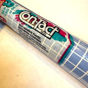 May include: Rolled-up Contact brand self-adhesive plastic decorative covering. The roll features a blue and white grid pattern with teal and magenta accents. Text on the label reads "Easy to use, easy to move during application."