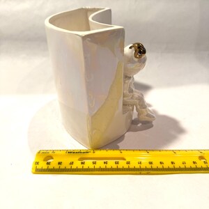 May include: A white ceramic vase with a pearlescent finish, featuring a seated astronaut figurine with a gold helmet. The vase is approximately 5 inches tall and has a unique, curved design. A yellow ruler is in the foreground.
