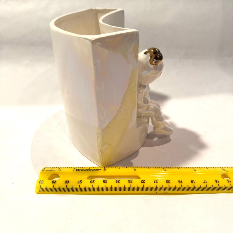 May include: A white ceramic vase with a pearlescent finish, featuring a seated astronaut figurine with a gold helmet. The vase is approximately 5 inches tall and has a unique, curved design. A yellow ruler is in the foreground.