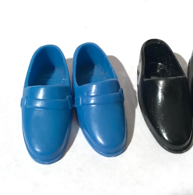 May include: A pair of blue and a pair of black plastic doll shoes. The shoes have a simple design with a strap across the top.