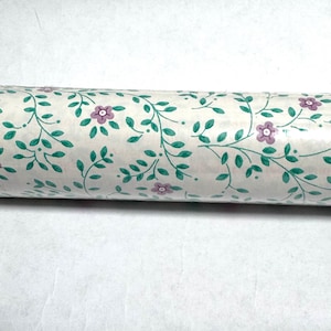 May include: Rolled wallpaper featuring a repeating pattern of green vines, leaves, and small purple flowers on a cream background. The wallpaper is rolled up, showing the detailed floral design.