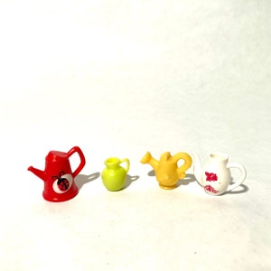 May include: Four miniature plastic watering cans in different colors and designs. A red watering can with a ladybug design, a yellow watering can, a green watering can, and a white watering can with a floral design.