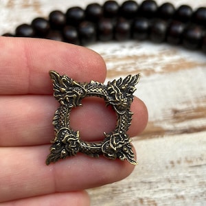 Pu&ograve; includere: Un piccolo ciondolo quadrato color ottone antico con un design floreale e di drago. Il ciondolo ha un'apertura circolare centrale ed &egrave; tenuto vicino a una collana di perline marrone scuro. Il ciondolo misura circa 2,5 cm quadrati.