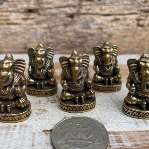 Op de afbeelding: Vijf kleine koperen Ganesha-beeldjes in zittende positie, elk met een olifantenkop, grote oren en gedetailleerde versieringen op de basis. Een Amerikaanse kwartje ligt op de voorgrond om de schaal van de beeldjes aan te geven.
