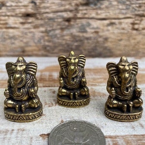 Op de afbeelding: Drie kleine, antiek ogende messing beeldjes van de godheid Ganesha, elk zittend afgebeeld met een olifantenkop en grote oren. De beeldjes staan in een rij opgesteld, met een muntstuk op de voorgrond voor een indicatie van de schaal.