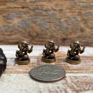 K&ouml;nnte beinhalten: Drei kleine, goldfarbene Ganesh-Statuen sind auf einer Holzoberfl&auml;che angeordnet. Eine M&uuml;nze befindet sich neben den Statuen. Ein dunkelbraunes Perlenarmband befindet sich auf der linken Seite des Bildes.