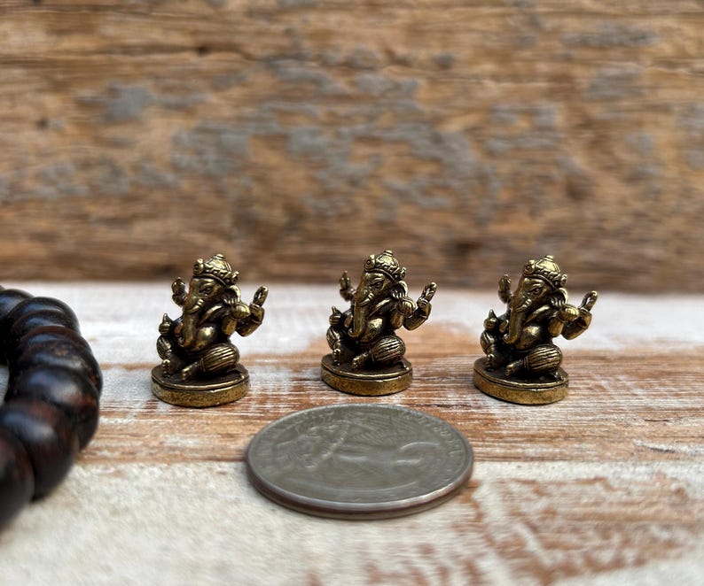 K&ouml;nnte beinhalten: Drei kleine, goldfarbene Ganesh-Statuen sind auf einer Holzoberfl&auml;che angeordnet. Eine M&uuml;nze befindet sich neben den Statuen. Ein dunkelbraunes Perlenarmband befindet sich auf der linken Seite des Bildes.