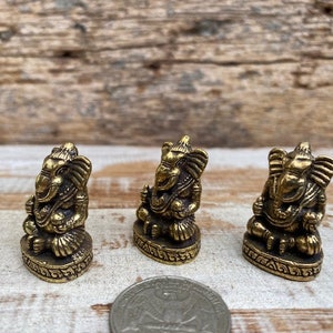 Op de afbeelding: Drie kleine koperen Ganesha-beeldjes in zittende houding, elk met gedetailleerde snijwerken van de slurf en oren, weergegeven naast een muntstuk om hun miniatuurformaat aan te duiden.