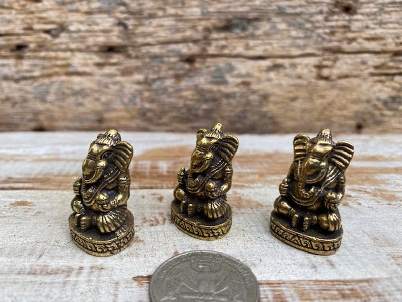 Op de afbeelding: Drie kleine koperen Ganesha-beeldjes in zittende houding, elk met gedetailleerde snijwerken van de slurf en oren, weergegeven naast een muntstuk om hun miniatuurformaat aan te duiden.